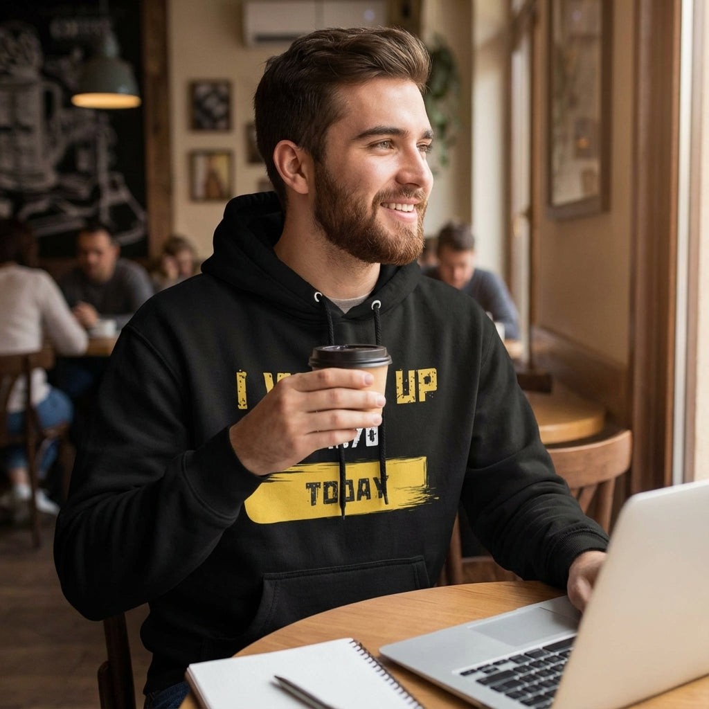 Man in a black hoodie with yellow text holding a coffee cup, sitting at a table with a laptop in a cafe.