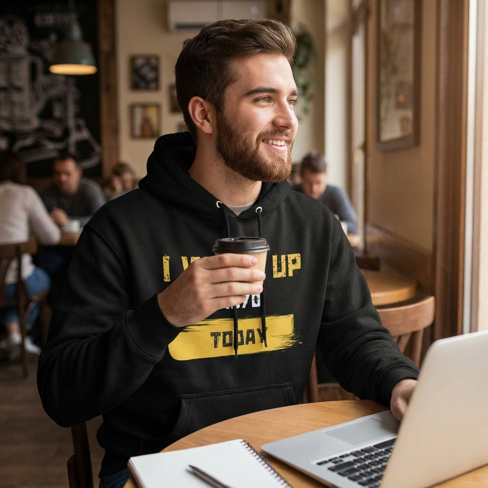 Man in a black hoodie with yellow text holding a coffee cup, sitting at a table with a laptop in a cafe.